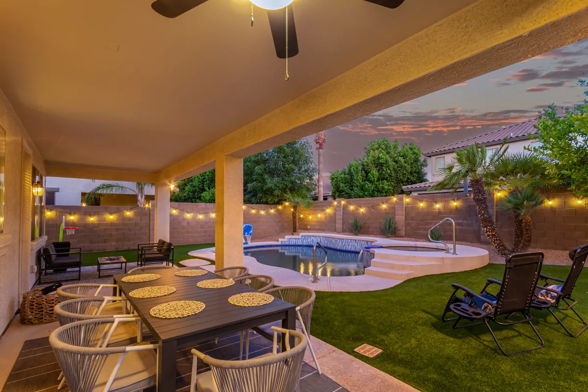 An inviting covered patio with a stylish dining table and chairs overlooks the swimming pool. Elegant landscape lighting and string lights create a warm ambiance, perfect for evening relaxation or alfresco meals. The sunset sky adds to the tranquil, vacation-like atmosphere.