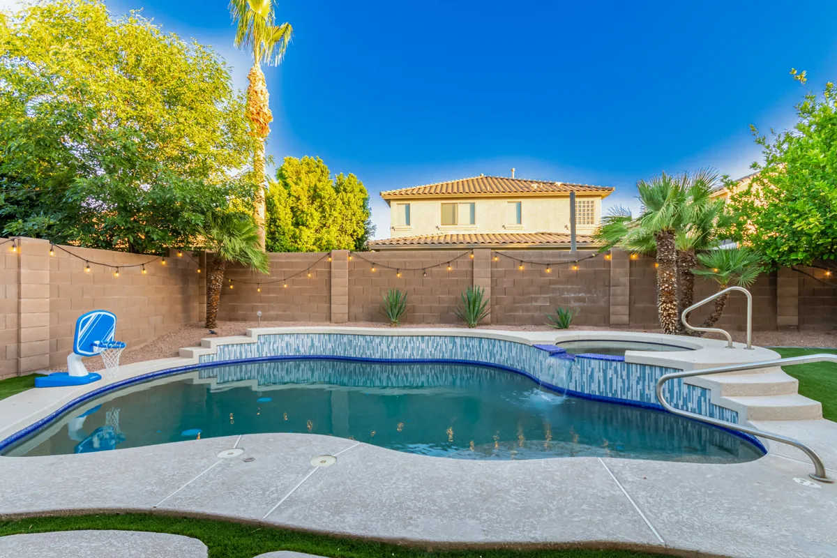 A vibrant backyard scene featuring a beautiful swimming pool, water features, lush landscaping, and a basketball hoop by the poolside. The area is sunlit and playful, offering spaces for both relaxing and active recreation.