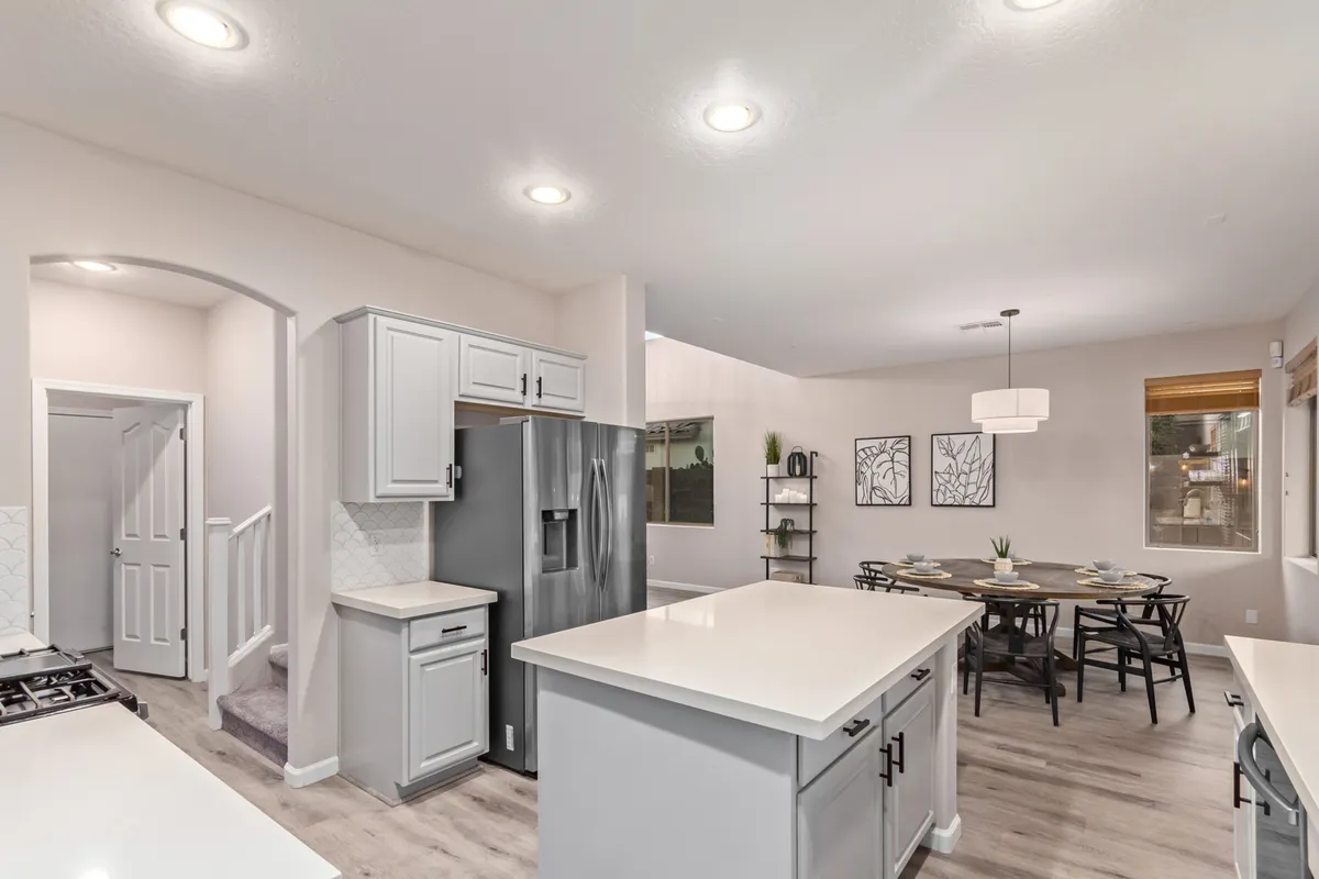 An inviting open-concept space is shown with a kitchen island at the forefront, a dining table nearby, and large windows flooding the room with natural light. The visible kitchen and dining spaces hint at celebrations, family meals, and effortless hosting, all set within a sunlit, warm environment.