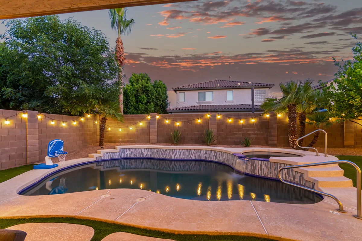 A spacious backyard showcases a sparkling swimming pool surrounded by comfortable patio seating and twinkling string lights. The landscaped area sets a festive, inviting mood perfect for group gatherings. A large patio area adjacent to the pool is ready for BBQs or lounging after a swim.