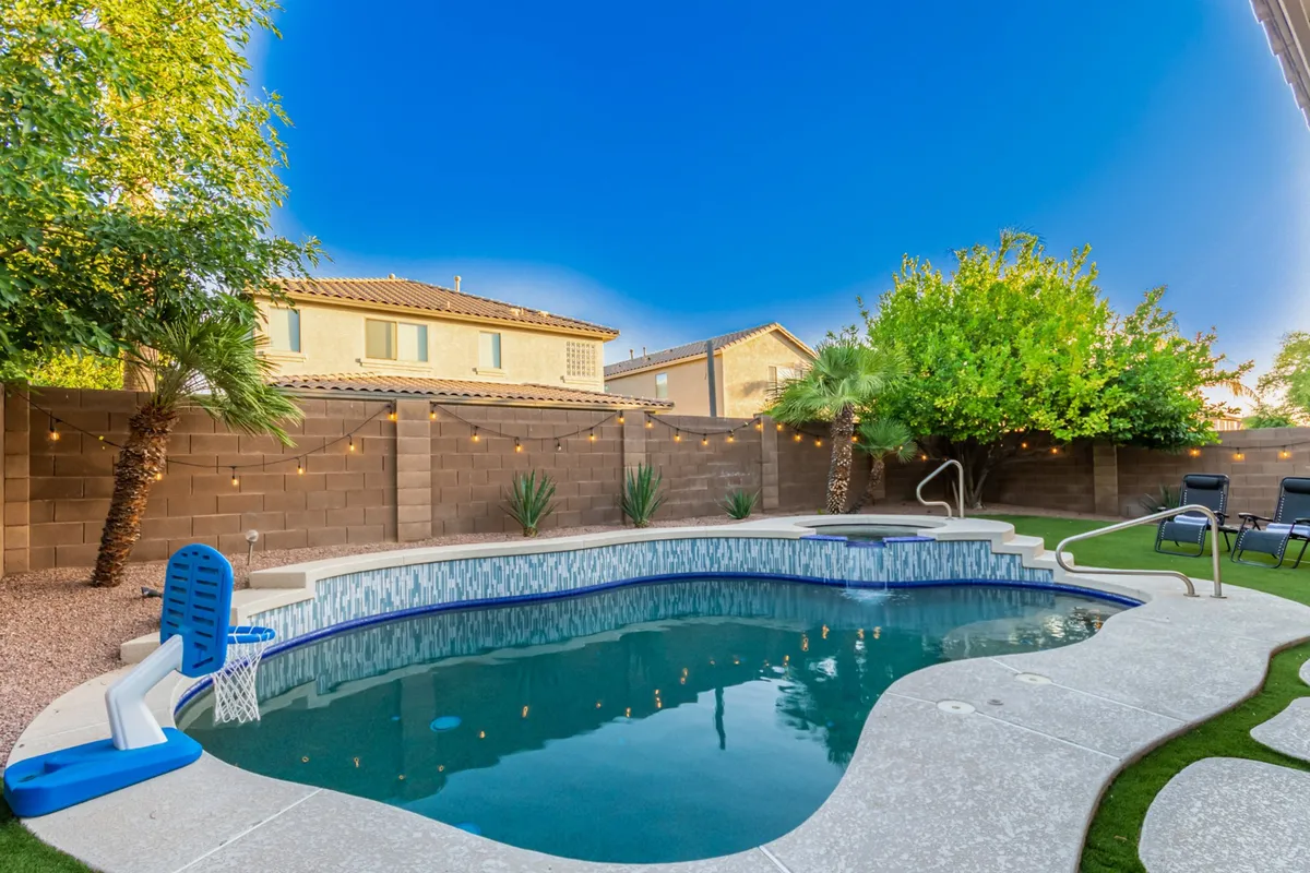This exterior image showcases a sparkling swimming pool surrounded by lush greenery and a backyard seating area adorned with string lights. The scene suggests a tranquil, residential setting ideal for unwinding or socializing outdoors.