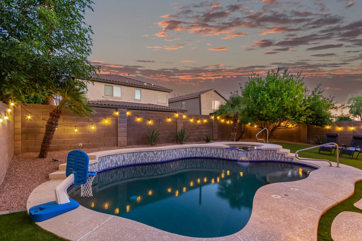 A wide view of the backyard with a shimmering swimming pool as the centerpiece, surrounded by lush grass and mature landscaping. String lights overhead hint at evening festivities, and the open lawn offers space for family games or playful gatherings.