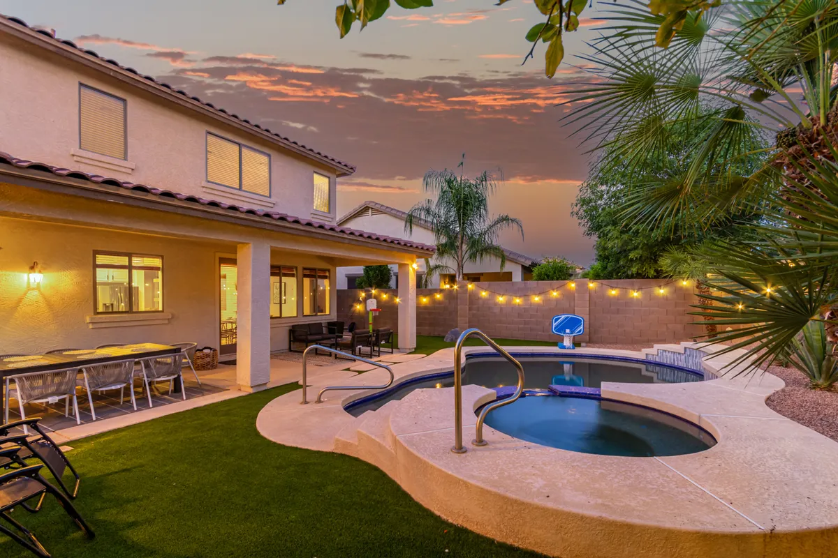 A backyard oasis featuring a sparkling swimming pool surrounded by lush greenery, patio seating, and festive string lights overhead. The space is set up for both relaxation and social gatherings, with areas for lounging, outdoor dining, and group activities.