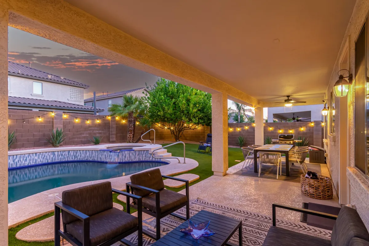 A twilight view of the backyard featuring a sparkling swimming pool, inviting outdoor seating area, string lights overhead, and a dining table perfect for alfresco meals. The setting is lively and cheerful, ideal for both daytime play and evening gatherings.