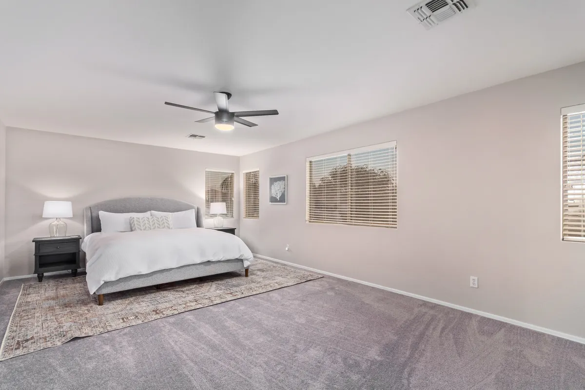 A tranquil bedroom featuring a cozy bed with layered bedding, a nightstand with a lamp, a soft rug underfoot, two windows bringing in natural light, and a ceiling fan overhead. The room exudes calm and is ideal for restful sleep or quiet reading.