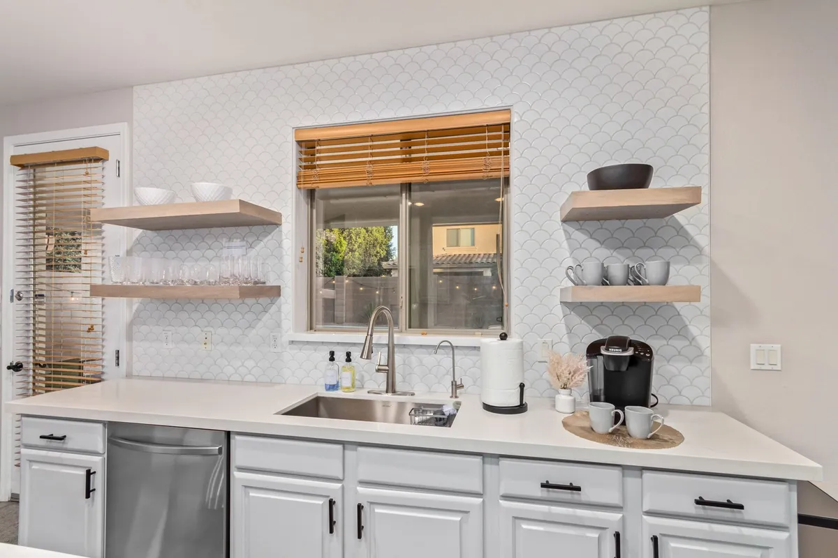 This interior image presents a modern kitchen with a sink, window with wooden blinds, shelves filled with cups and glasses, a coffee maker, and tasteful decorative items. The space feels inviting and perfectly suited for meal preparation and daily routines.